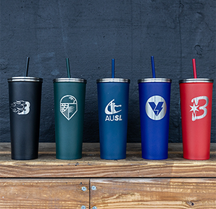 AUSL tumblers on a wooden dugout bench. From left to right: black tumbler with silver Blaze logo, green tumbler with silver Talons logo, navy tumbler with silver AUSL figure mark, purple tumbler with silver Volts logo, and red tumbler with silver Bandits logo.