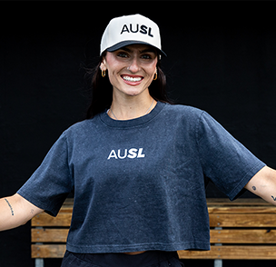 Baylee Klingler smiles wearing a black AUSL Cropped Tee with a white AUSL wordmark across the chest and a white cap with black AUSL wordmark.