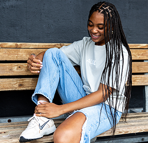 Athlete sits on a wooden dugout bench with a beige tee that says AUSL in white font across the chest, sporting ripped jeans and Nike sneakers.