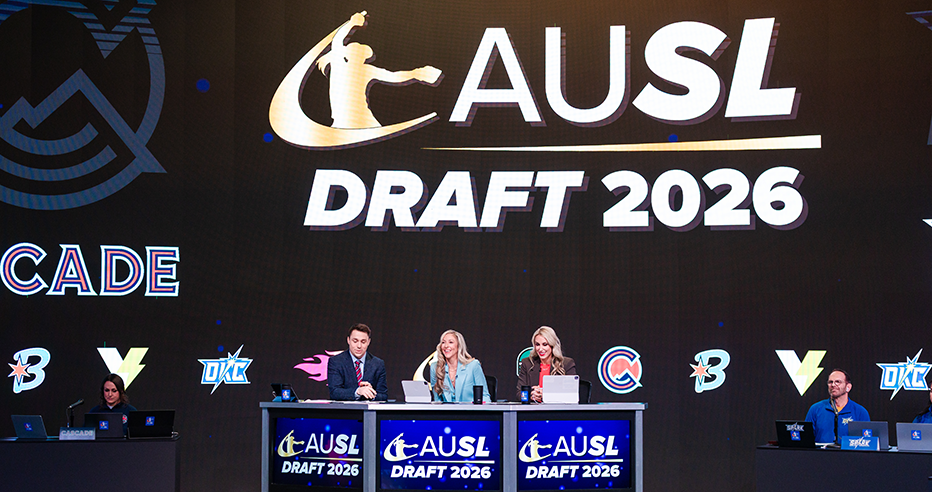 Broadcasters Chuckie Kempf, Amanda Scarborough, and Danielle Lawrie seated at the desk on the 2026 AUSL Draft set with a large black screen featuring the six team logos behind them.